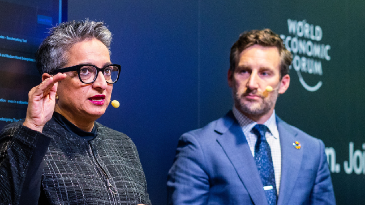 Close-up of two speakers, one looks at the other, both wearing headset microphones. 
