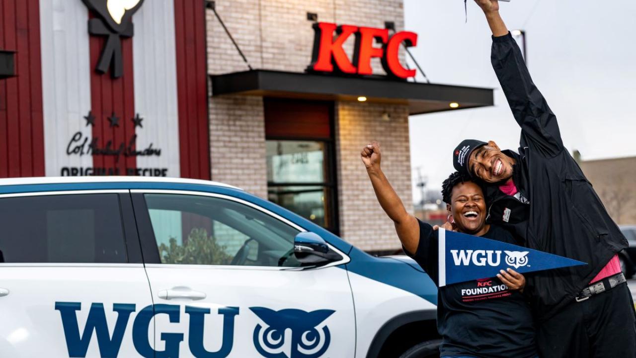 Two people smiling with one arm raised. One holds a WGU pendant. Standing in front of a car with WGU and logo on the side and all in front of a KFC restaurant.