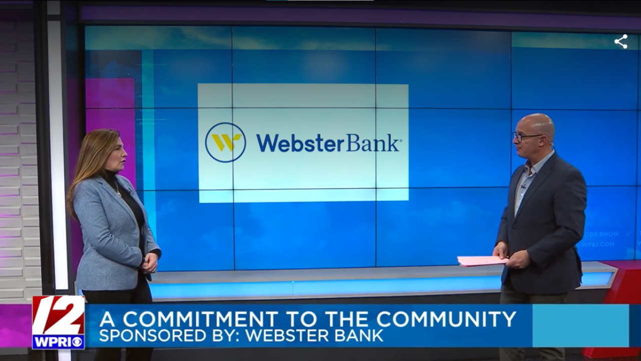 Two people standing in front of a large digital screen, one with papers, the other being interviewed.