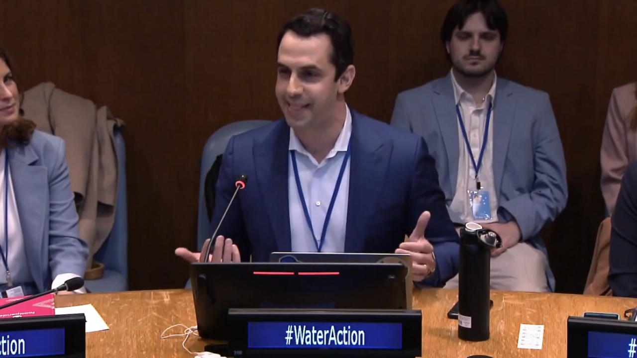 A person seated at a circular table with microphone and placard "#wateraction" Others to their sides and seated behind them.