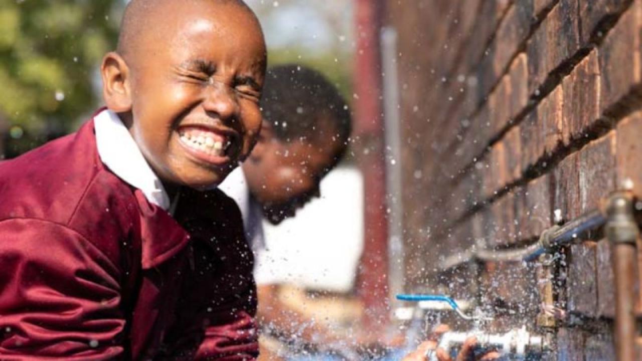 A smiling child splashing in an outdoor faucet.