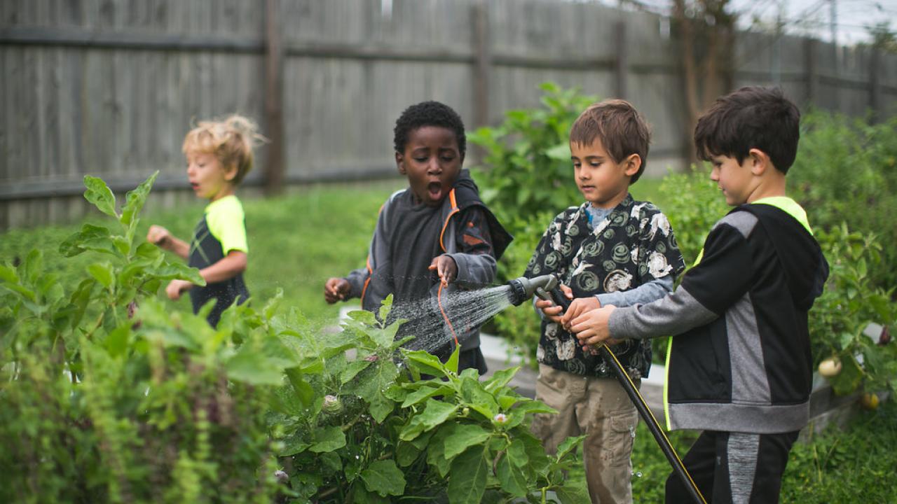 Children in a garden, one using a hose to water plants.