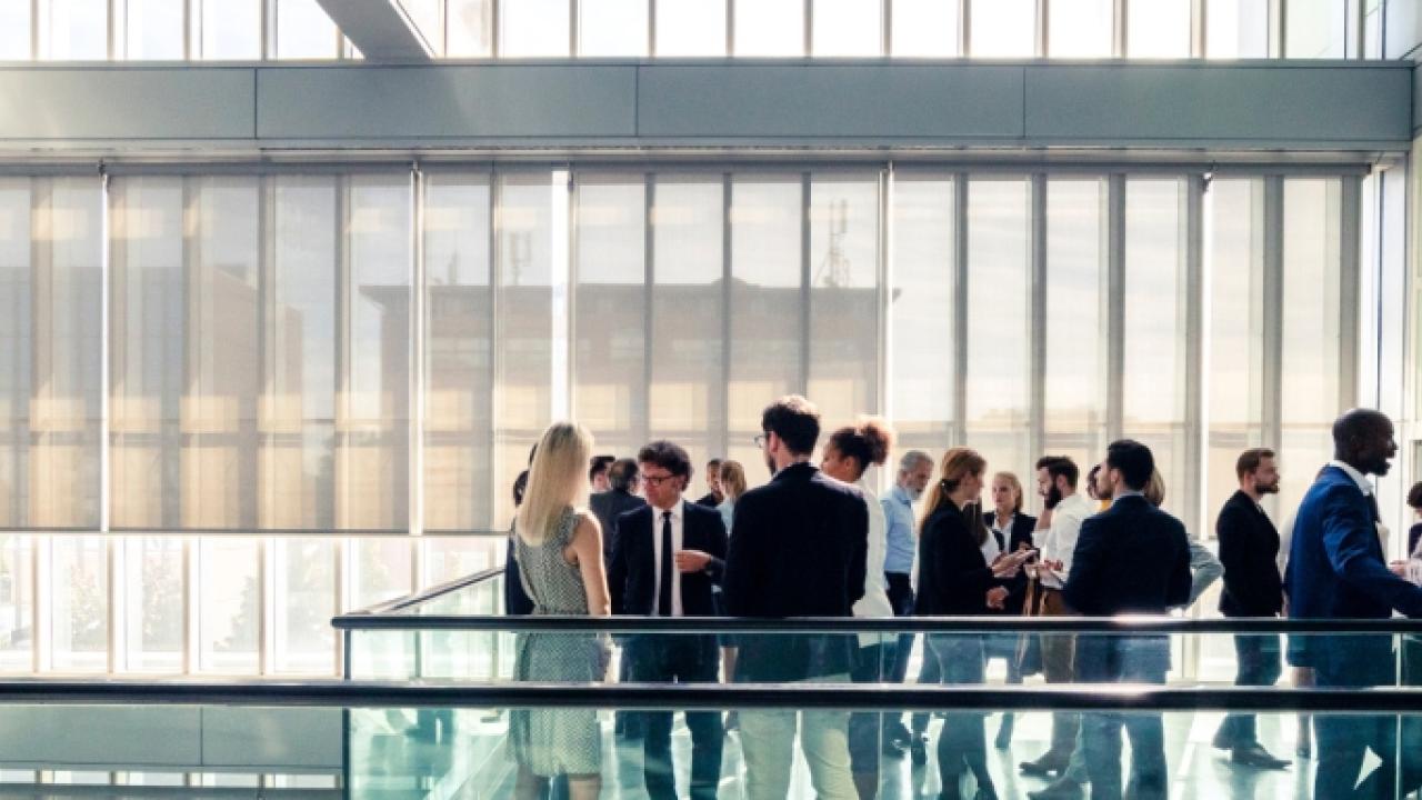 Group of about 25 business-attired people stand in small groups on a balcony in an office building with lots of outside light through glass windows