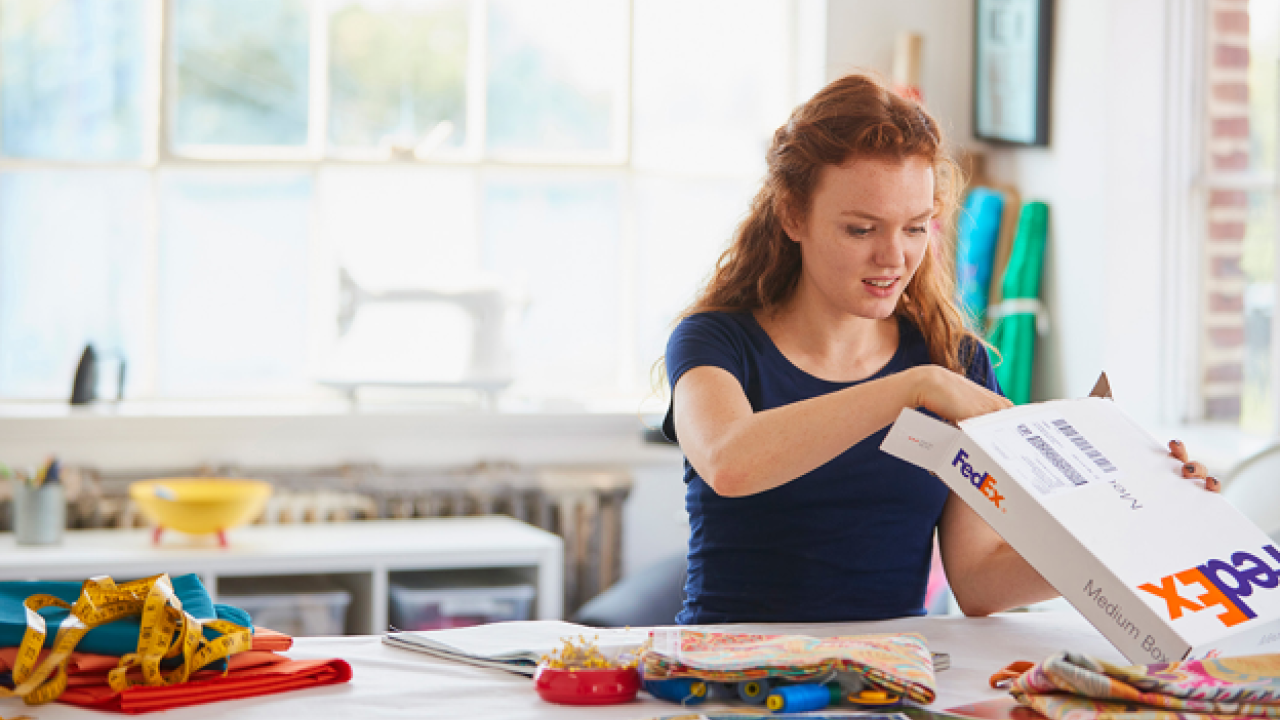 a person in a bright room, sitting at a large table with craft items, opening a FedEx box