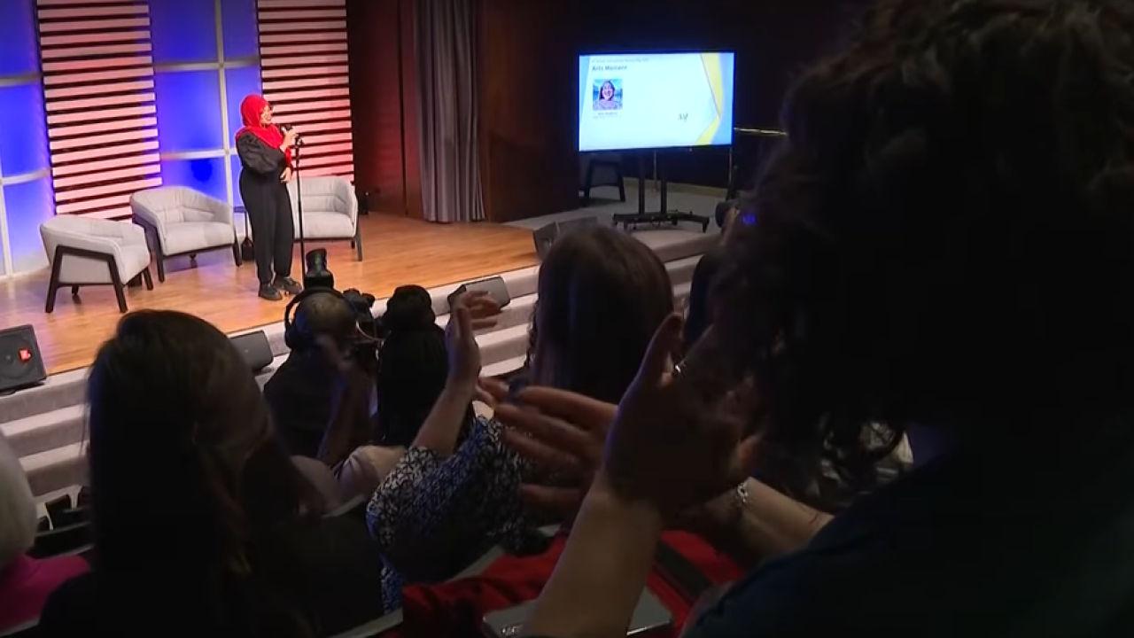 Saleemah Abdul-Ghafur speaking on a stage with three armchairs. An audience in front of her.