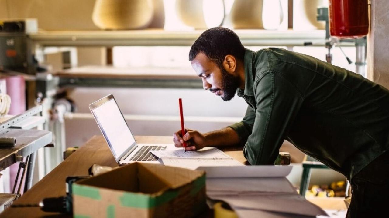 A person leant on a desk writing in a book that it resting on a laptop
