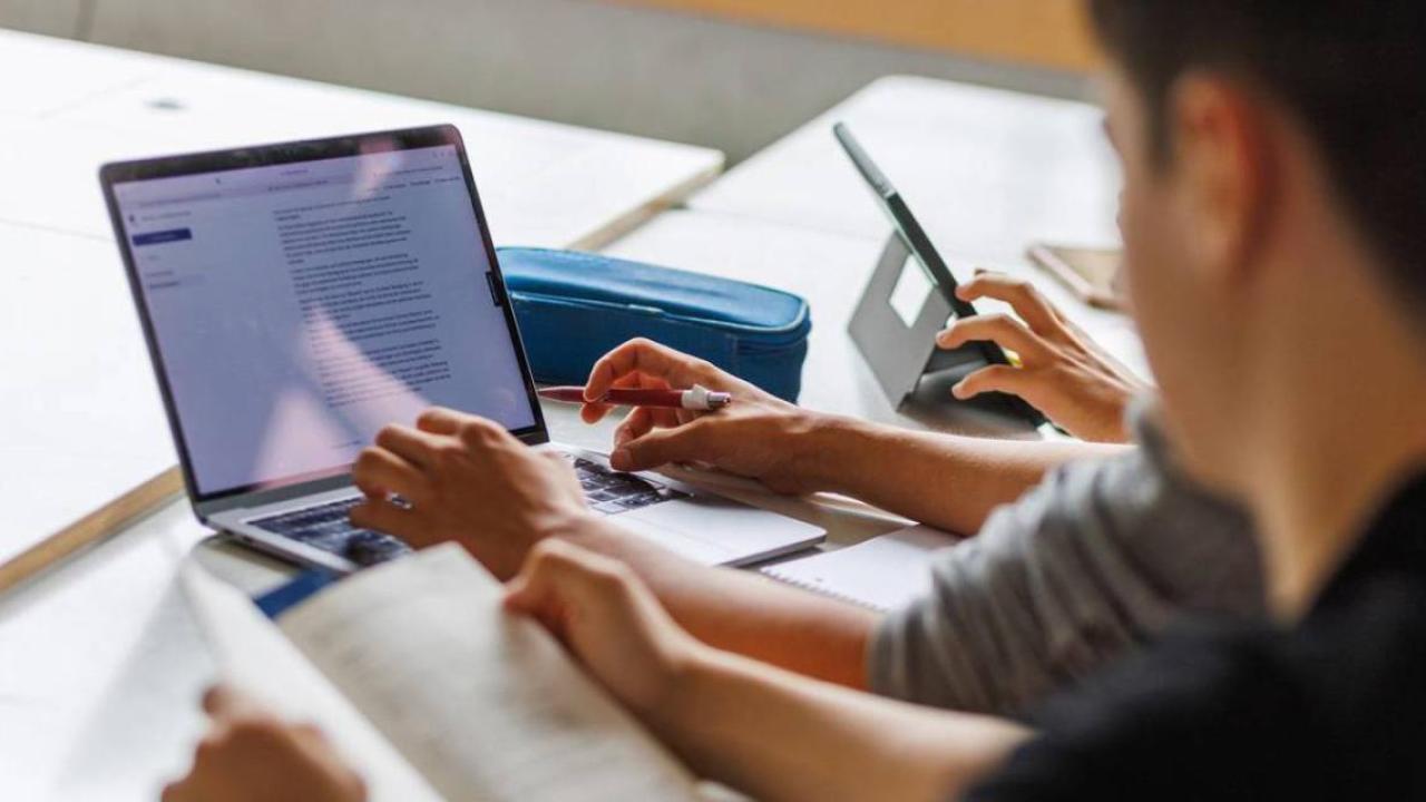 Looking over the shoulder of a person with an open book, another to their right using a laptop on a desk.