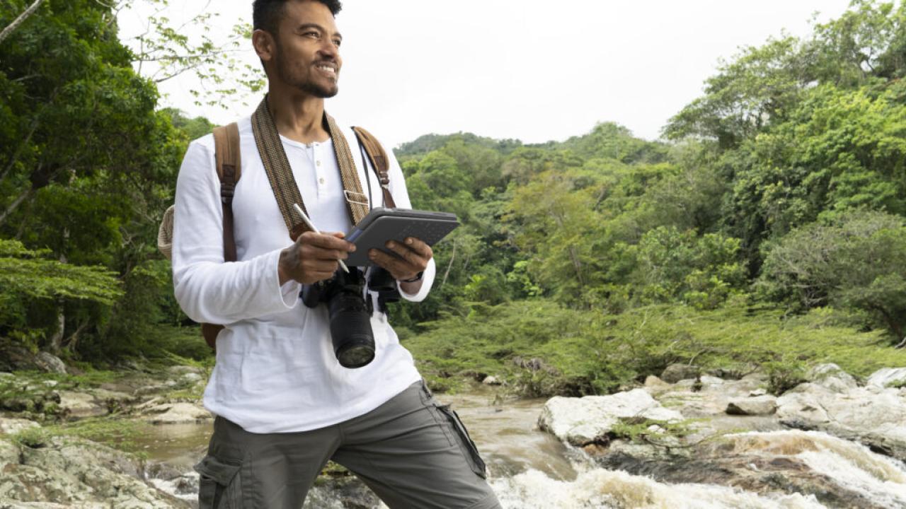 A smiling person with a camera and notepad next to a stream