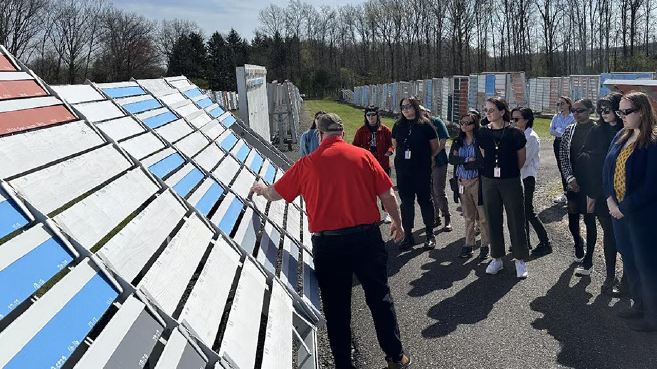 A person speaking to a group outside, a rack of painted boards to the side.