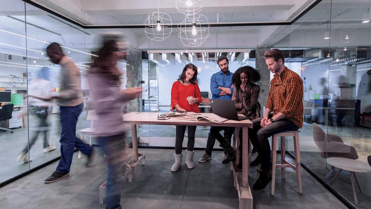 Four people looking at a laptop on a tall desk. Blurred others going by in a busy office setting.