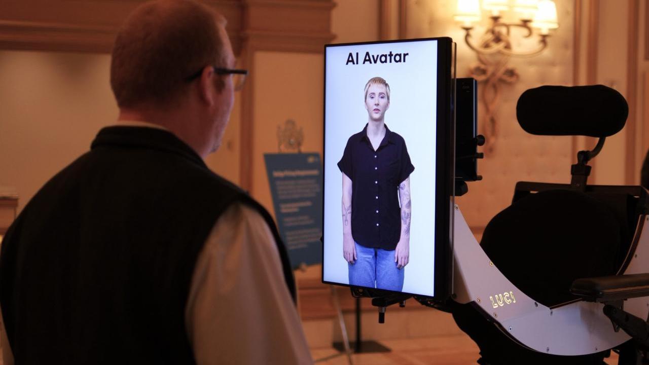 Erin Taylor appearing as an avatar on a small screen, another looks at it.