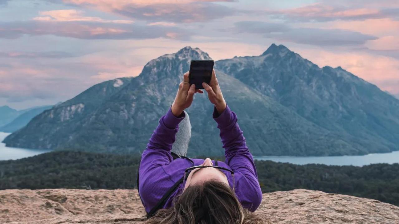 A person laying on a rock looking at their phone held above them. A mountain landscape in the distance.