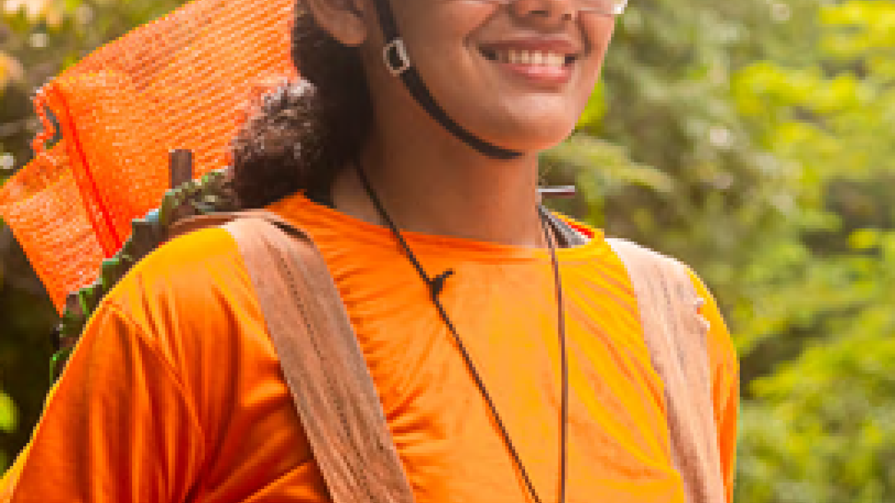 A smiling person in bright orange and safety gear in a forest setting.