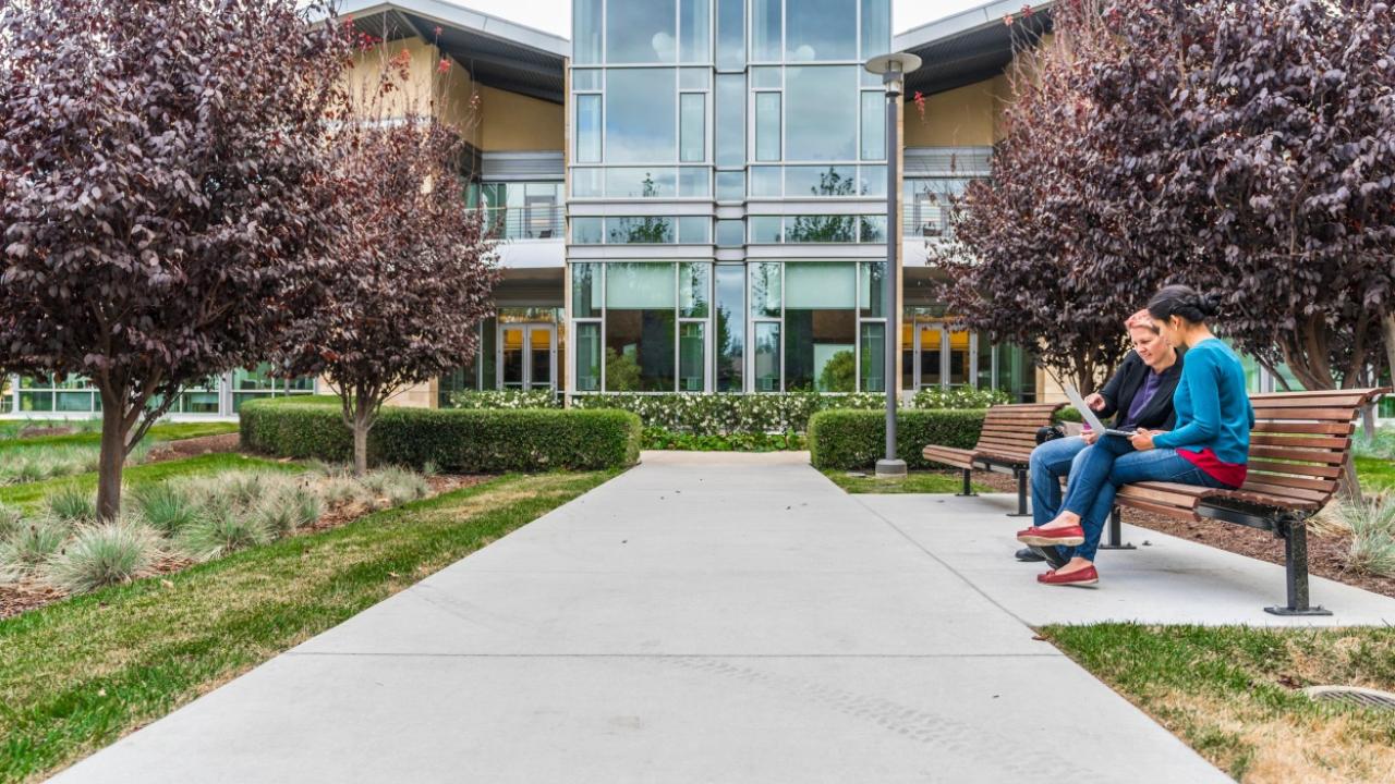 Two people sitting on a bench outside a building in a park like setting.