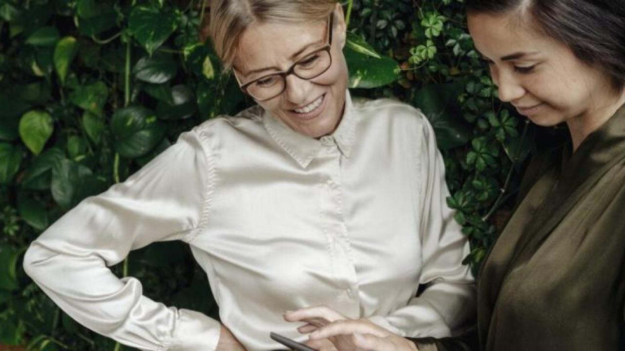 Two people looking at something together and smiling, in front of greenery