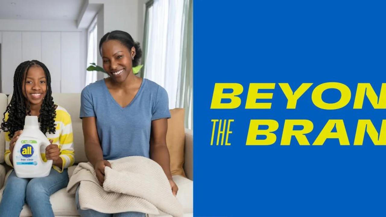 An adult and child sitting next to each other on a couch. One folding laundry and the other holding a bottle of detergent.