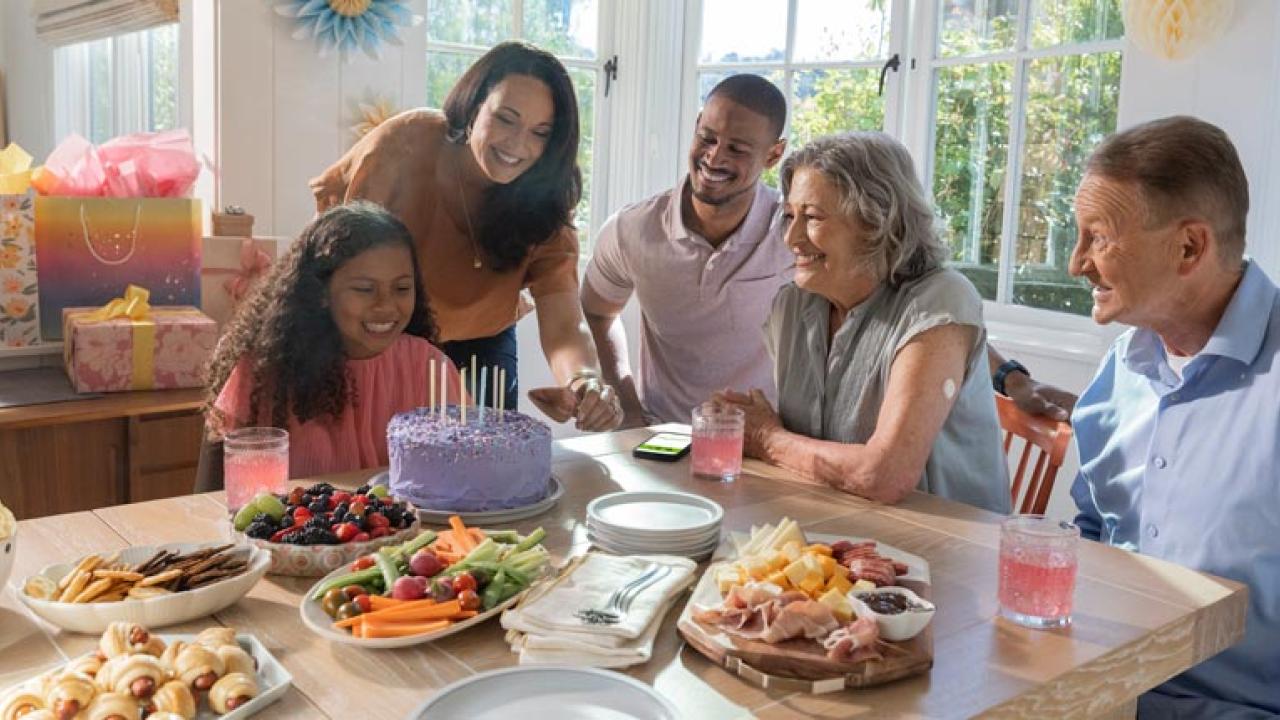 People sat around a buffet table, looking at a person who is sat in front of a celebratation cake 