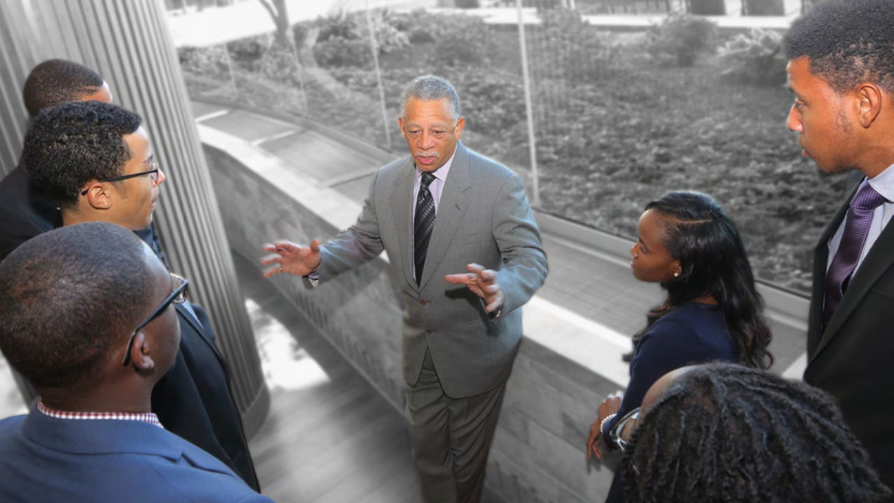 a group of people gather around one speaker in front of a wall of windows