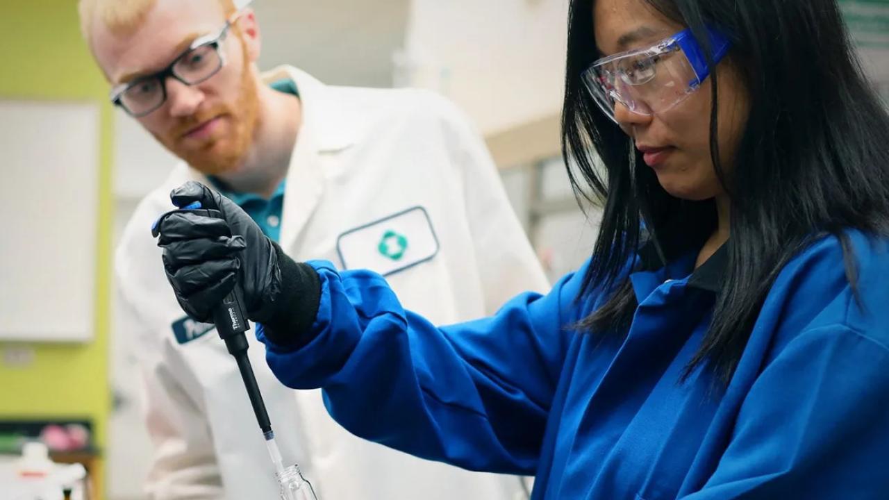 Two chemists, one observing another using a pipette.