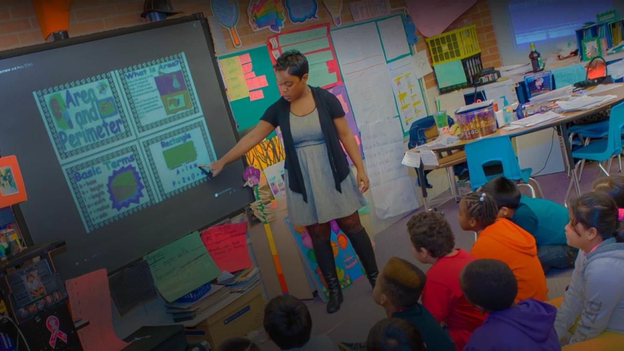 A teacher in front of a class of students, looking at a screen with lesson in area and perimeter on it.
