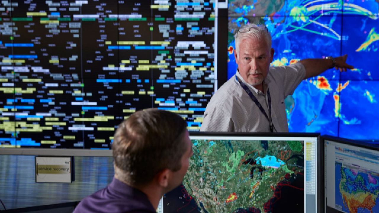 in a room with large screens on the walls, a person points at a global map while talking to a seated person looking at a computer screen with a map of the US