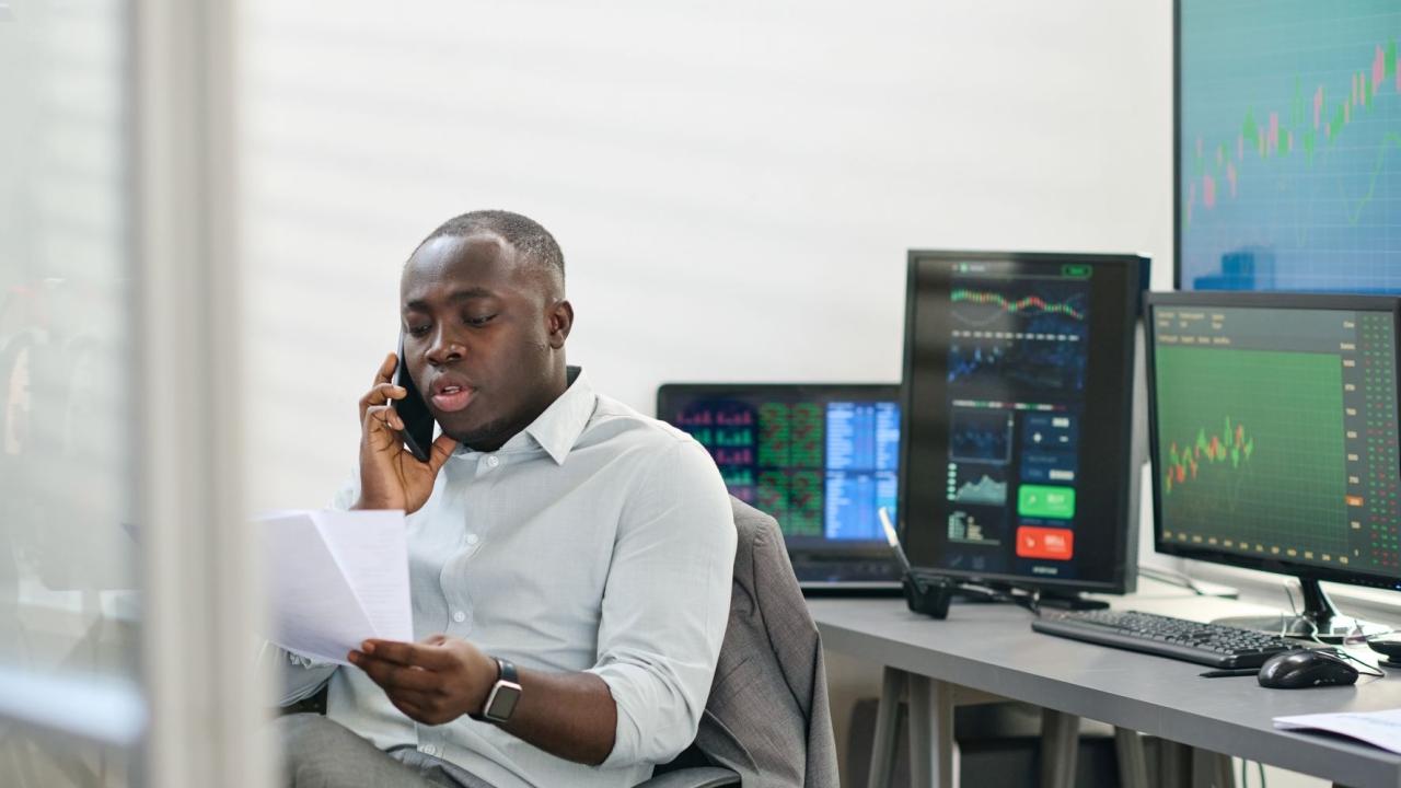 A person talking on a phone, looking at papers, their back to multiple computer screens.