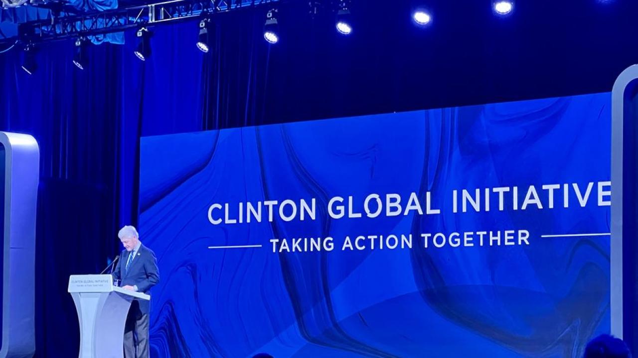 Bill Clinton at a podium, a large screen with "Clinton Global Initiative" behind him