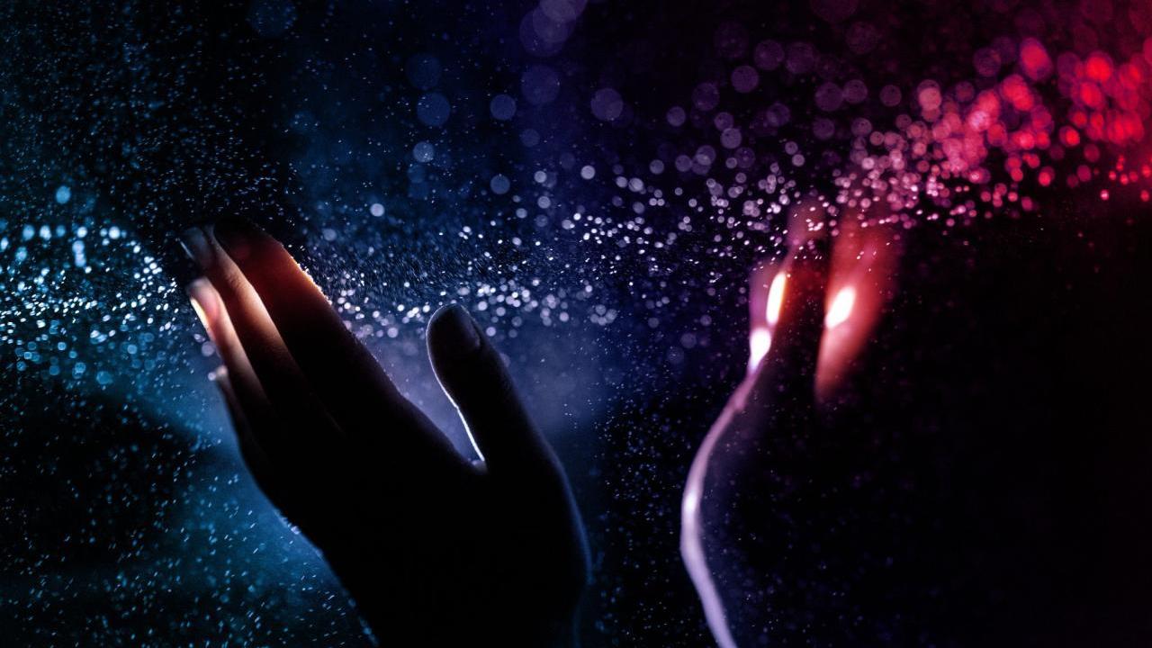 Two hands upheld in a dark space. A narrow band of colorful light and particles streaming across the finger tips.