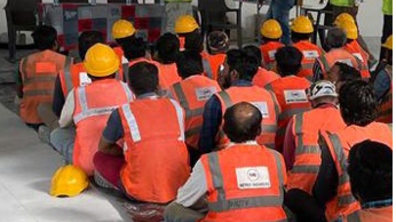 A group seated on the floor, each wearing high-vis vests and some in hard hats. People at the front of the room speaking to them.