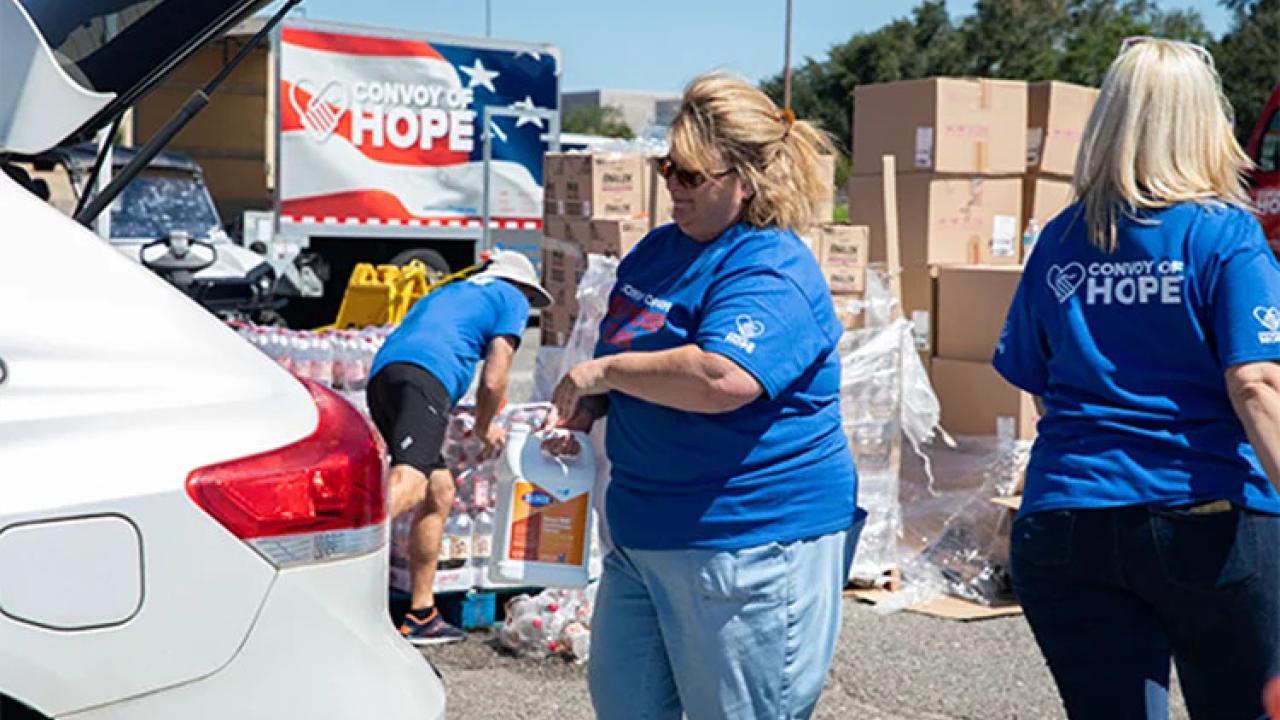 Volunteers loading the trunk of a vehicle. "Convoy of Hope" sign behind them and stacks of boxes.