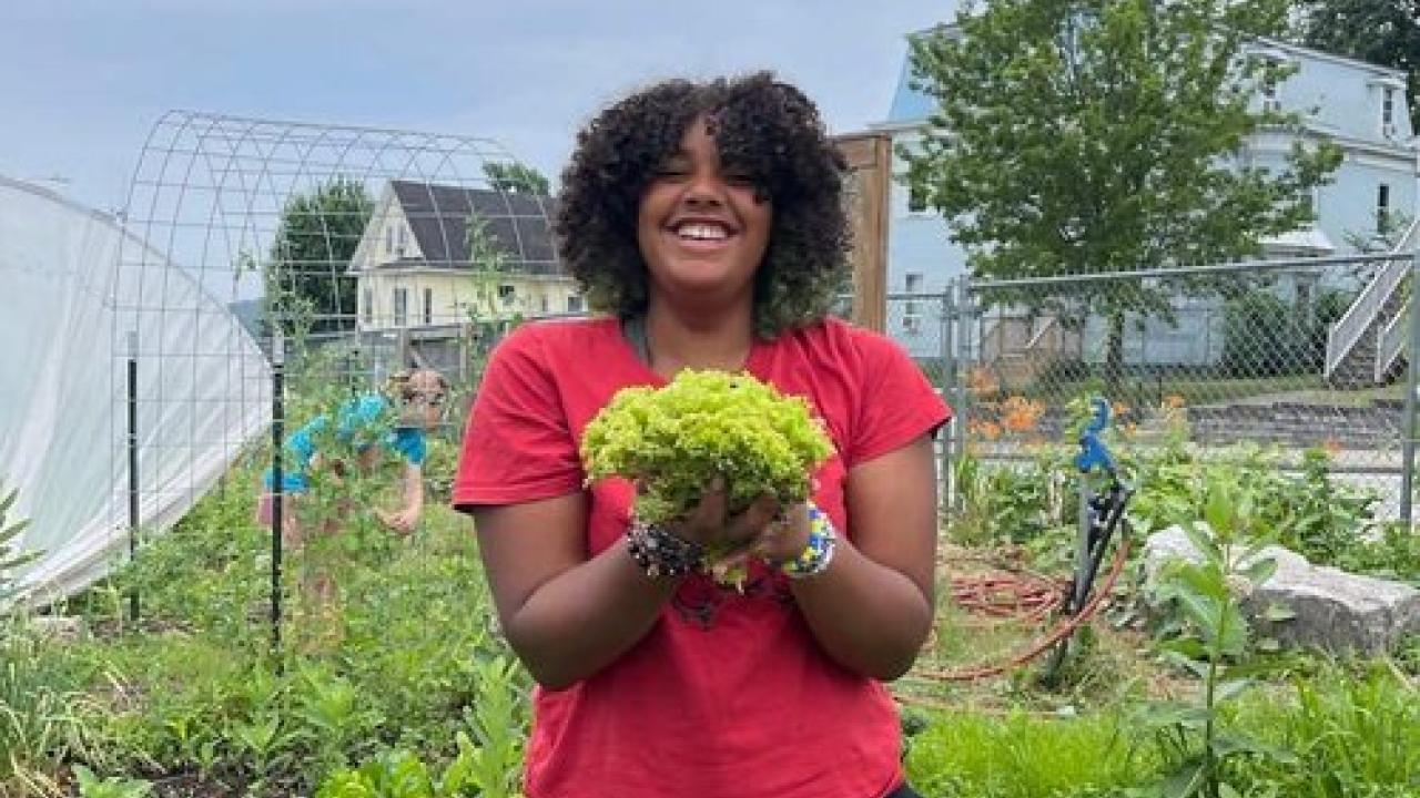 A smiling person holding a bundle of produce outside in a garden.