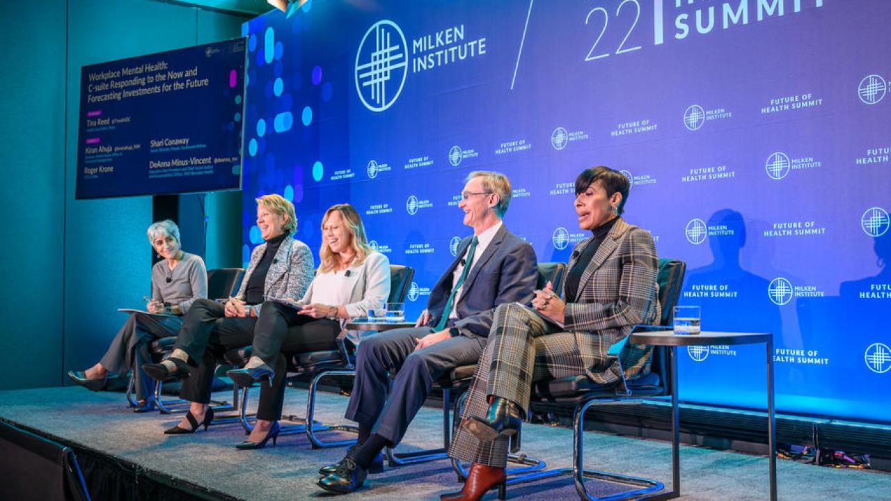 Five people seated on a small stage. A large display behind them "2022 Future of Health Summit" and Milken Institute logos.
