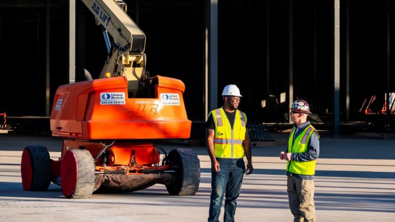 Two workers in hard hats and high vis vests stand in front of a large lift