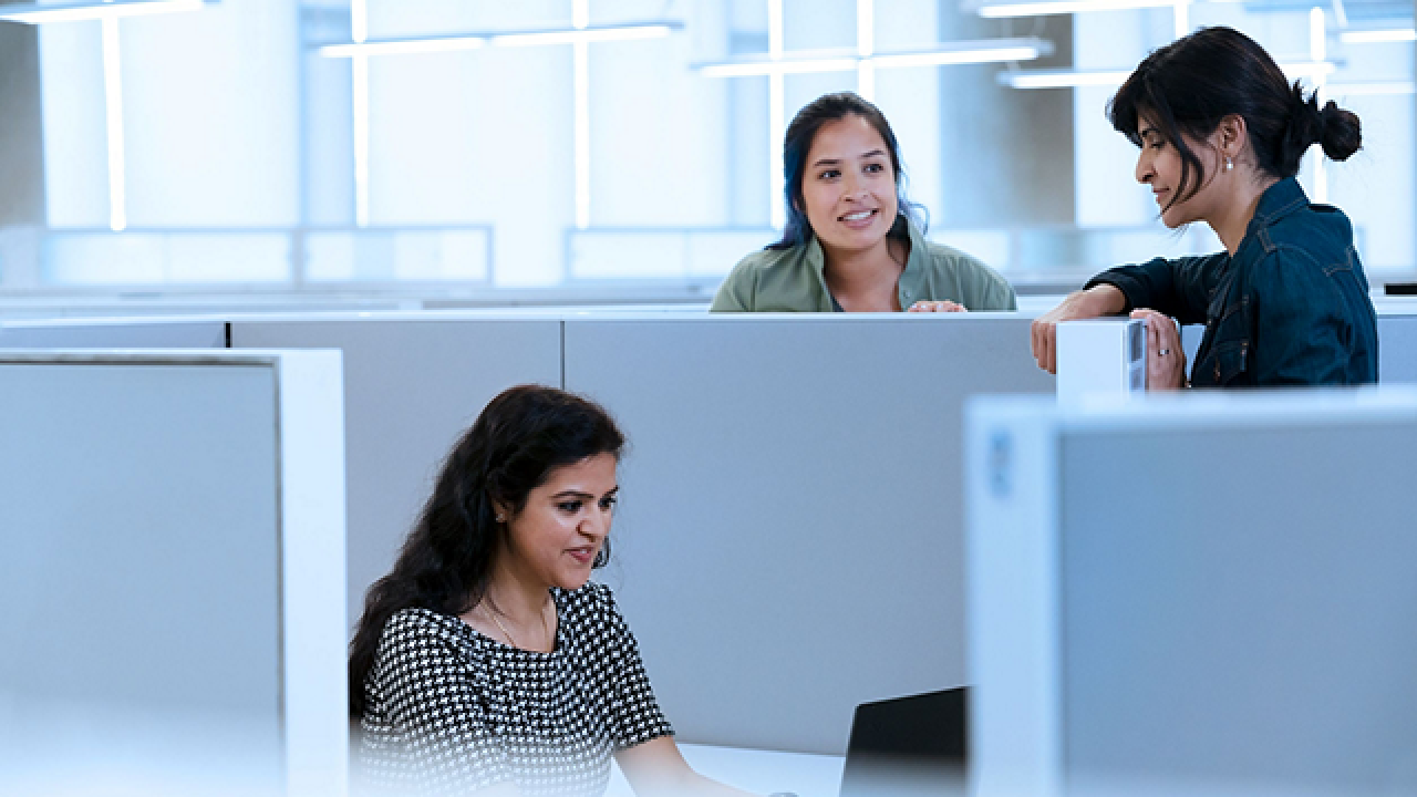 Two people standing, talking with a third who is seated at a desk in a cubicle, working on a laptop.