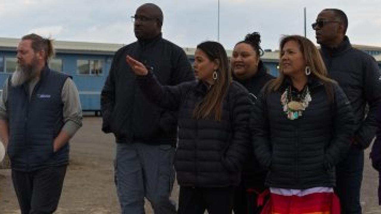 A group gathered outside looking in the same direction. One pointing. A blue long building behind them.