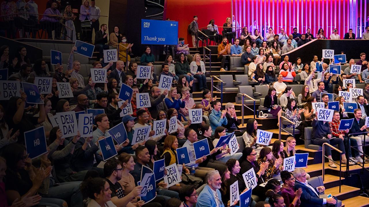 Crowd sitting in a theater at DE&I Day holding signs