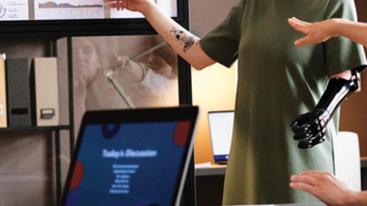 a person with a prosthetic left arm standing, pointing at a posted graph. Another sitting at a close table gesturing. "DEI Best place to work for disability inclusion 2022, 100% disability equality index" at the bottom