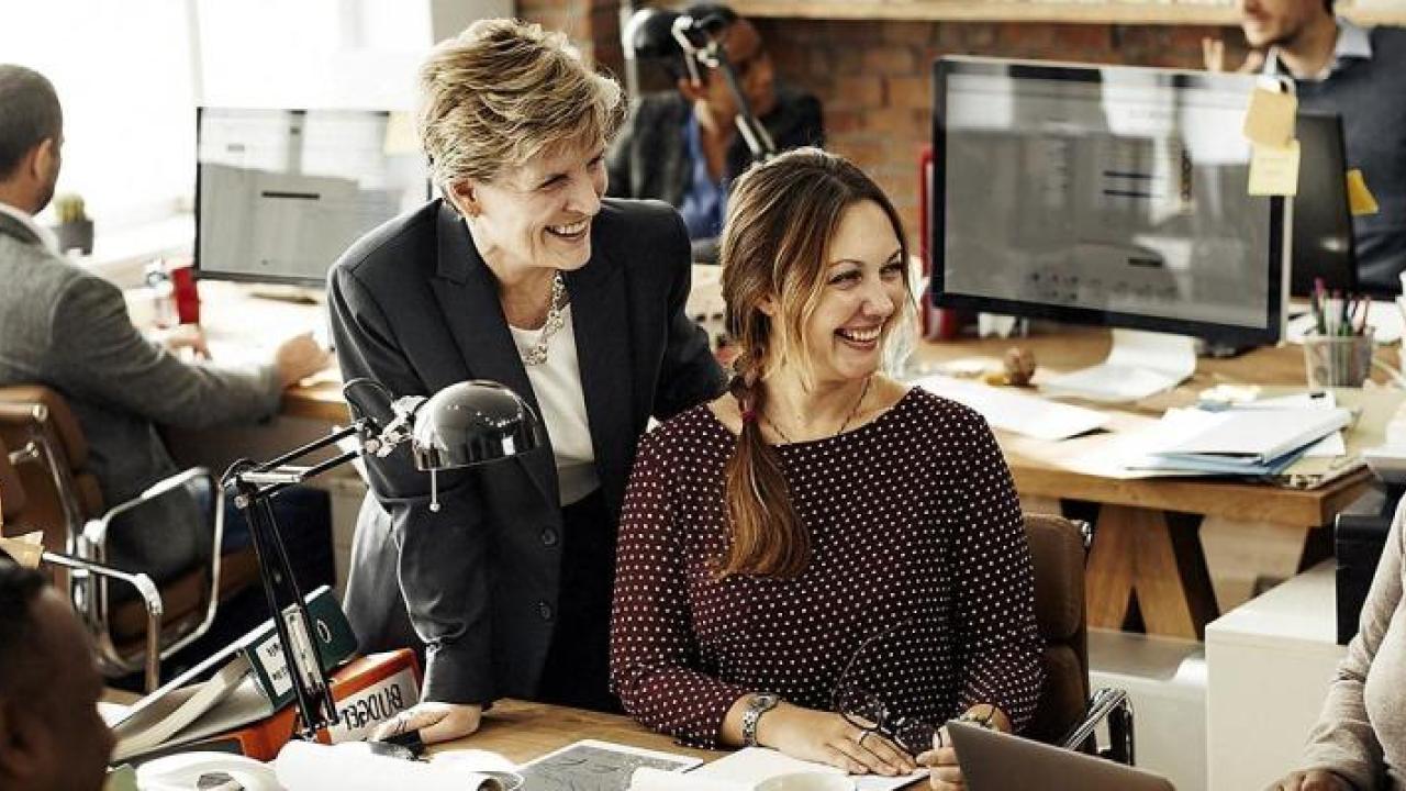 People in an office setting, three smiling at each other, sitting at a long desk, another at a computer behind them.