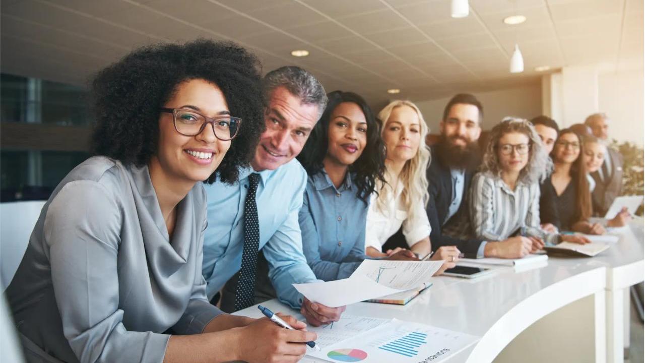 A row of diverse professionals at a long desk all looking at the camera.