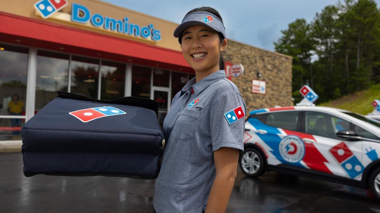 A smiling person in Domino's uniform, holding a pizza delivery box. A delivery car and Domino's store behind them.