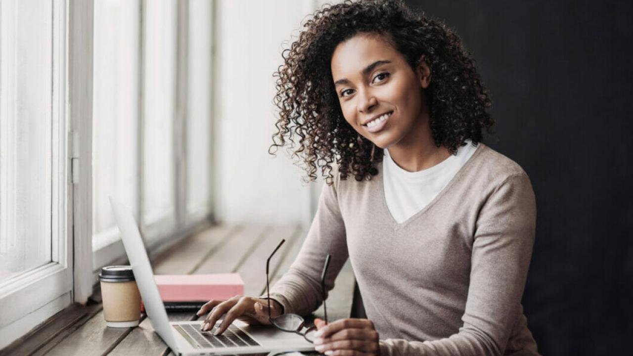 Young woman sitting at a desk, looking at the camera and typing on her laptop.