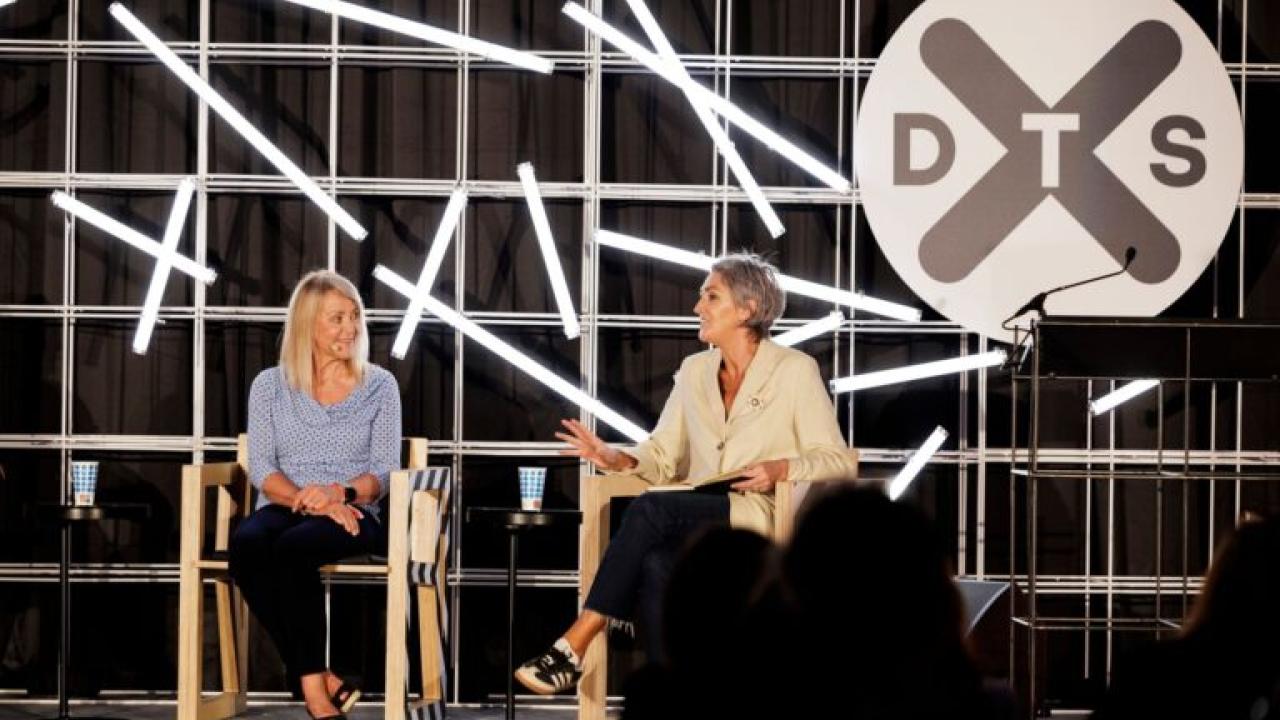 Two people seated on a stage. DTS logo displayed behind them.