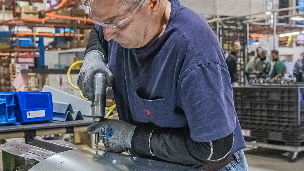 A worker in safety glasses and gloves drilling through a curved metal piece.