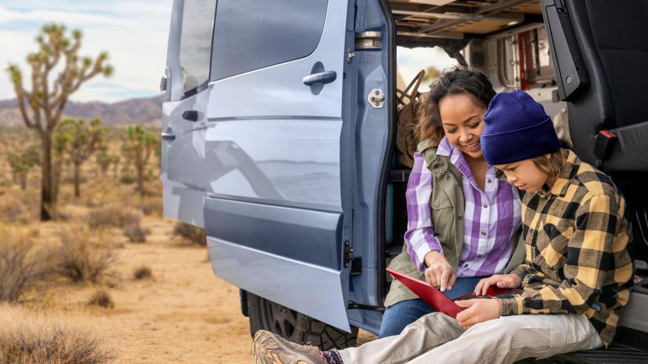 An adult and hild looking at an electronic tablet while sitting in the open side of a van in an arid area.