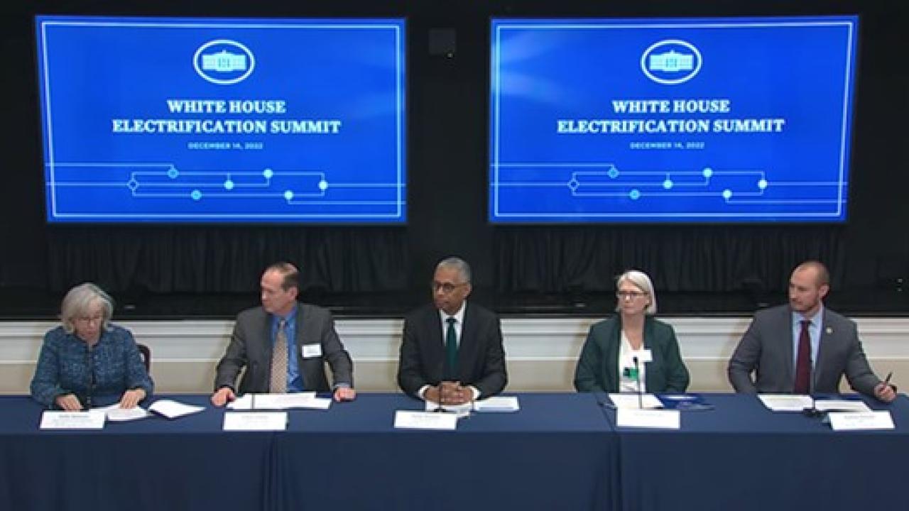 Five people sitting at a long desk, Two screens behind them with "White house Electrification summit" on them.