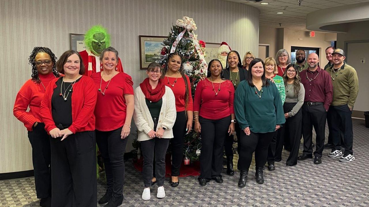 A group of volunteers posed in front of a chistmas tree. One dressed as the grinch behind them.