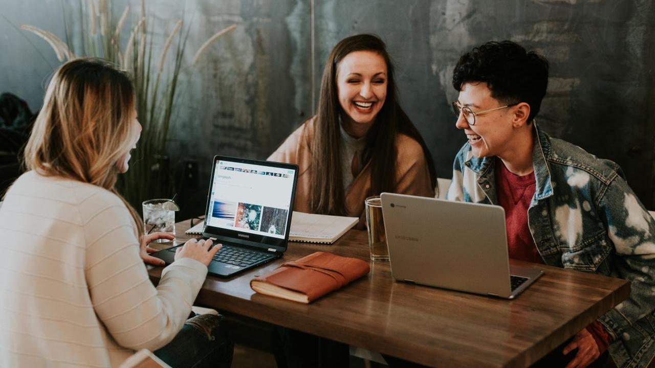 Three people seated at a shared table with laptops open, smiling.