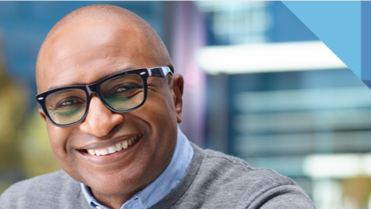 A person smiling, wearing black rimmed glasses. Blurred background.