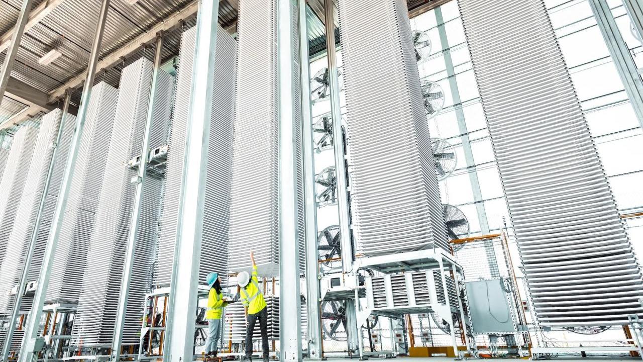 Two people in high-vis vests looking at a tall stack of metal sheets in a row of other stacks.