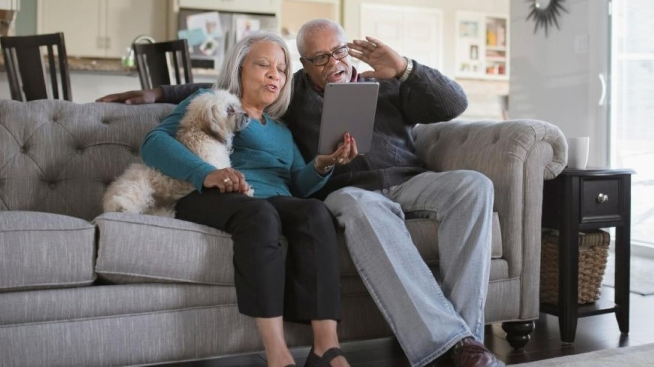 Two people and a small white dog sitting on a couch, looking at a tablet device.